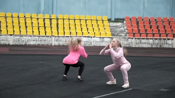 Kaukasische identieke mooie tweeling in sportkleding doen hurken en sprongen in het stadion — Stockvideo