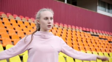 Spor giyimli genç bir bayan spor stadyumunun arka planında kollarını esnetiyor.