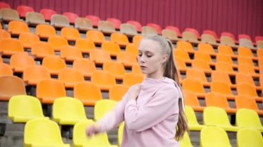 Spor giyimli genç bir bayan spor stadyumunun arka planında kollarını esnetiyor.