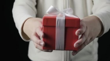 Küçük kızın ellerine yakın plan beyaz kurdeleli kırmızı kutuyu veriyor. Kış tatili, Noel, yeni yıl, sürpriz. Küçük çocuk siyah arka plandaki kutuda hediye veriyor. Doğum günü hediyesi olan kişi.