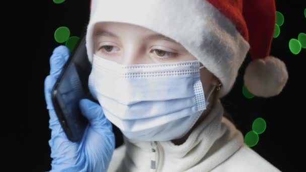 Девушка в шляпе Санта-Клауса, медицинская защитная маска, перчатки говорит по мобильному телефону — стоковое видео