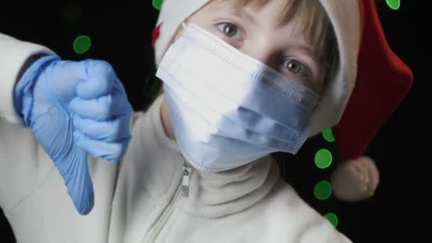 Девушка в шляпе Санта-Клауса, голубые перчатки, медицинская защитная маска показывает большой палец вниз — стоковое видео