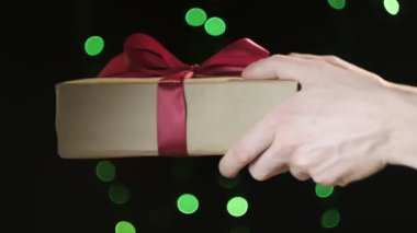 Eller, yeşil ışık saçan Bokeh 'in kırmızı kurdeleli Noel hediyesi kutusunu tutuyor.