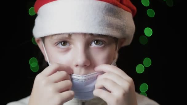 Niña en el sombrero rojo de Santa Claus se pone la máscara protectora médica, guantes azules — Vídeos de Stock