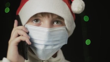 Kırmızı Noel Baba şapkalı, mavi tıbbi maskeli kız cep telefonundan konuşuyor.