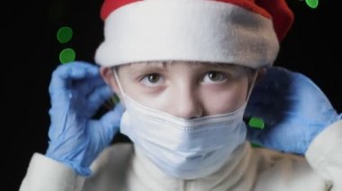 Kırmızı Noel Baba şapkalı, mavi eldivenli küçük kız tıbbi koruyucu maske takıyor.
