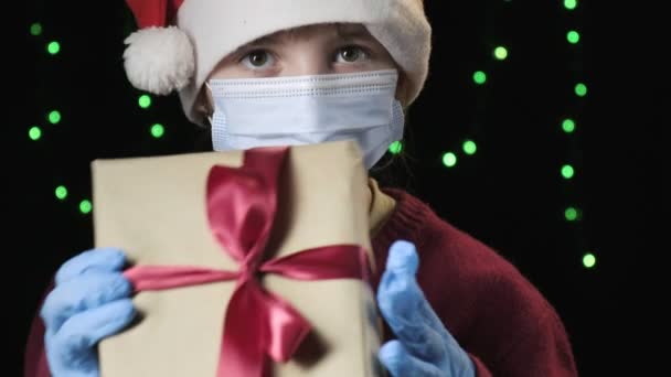 Girl in Santa Claus hat, medical protective mask, gloves holds gift with ribbon — Stockvideo