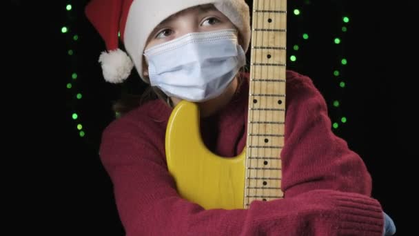 Fille dans le chapeau du Père Noël, masque de protection médicale bleu, gants tient la guitare — Video