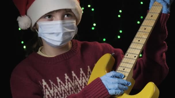 Κορίτσι στον Άγιο Βασίλη καπέλο, μπλε ιατρική προστατευτική μάσκα, γάντια κρατά κιθάρα — Αρχείο Βίντεο