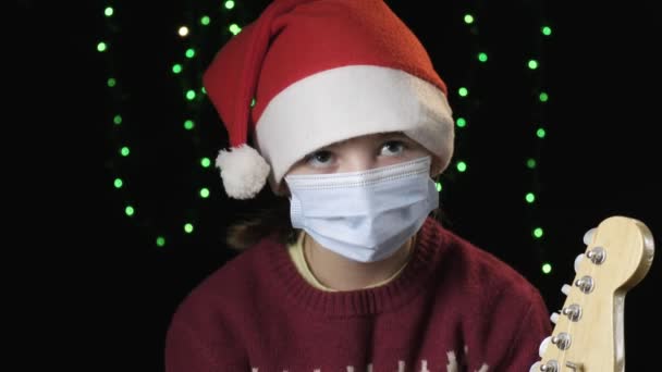 Dziewczyna w kapeluszu Świętego Mikołaja, niebieski medyczna maska ochronna, rękawiczki trzyma na gitarze — Wideo stockowe