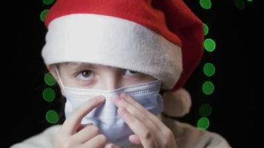 Kırmızı Noel Baba şapkalı küçük kız tıbbi koruyucu maske takıyor, mavi eldiven takıyor.