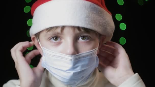 Маленькая девочка в красной шляпе Санта-Клауса надевает медицинскую защитную маску, голубые перчатки — стоковое видео