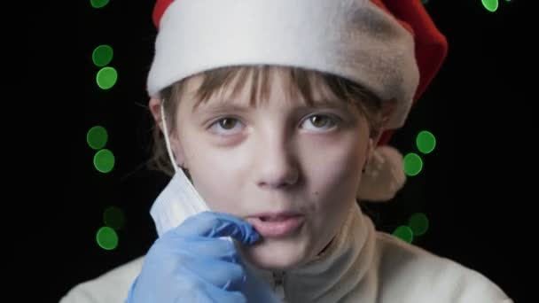Маленькая девочка в красной шляпе Санта-Клауса, голубые перчатки надевает медицинскую защитную маску — стоковое видео
