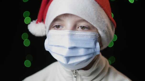 Boos meisje in rood kerstman hoed, blauw medisch beschermend masker kijkt naar camera — Stockvideo