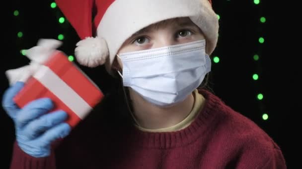 Girl in Santa Claus hat, medical protective mask, gloves holds gift with ribbon — Vídeos de Stock