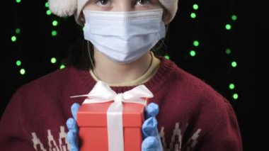 girl in Santa Claus hat, medical protective mask, gloves holds gift with ribbon