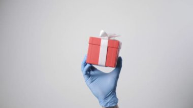 Elinde mavi koruyucu eldiven, elinde beyaz kurdeleli kırmızı hediye kutusu var. Noel, yeni yıl, doğum günü, Sevgililer Günü sürprizi. Beyaz arkaplanda küçük bir kutu bulunduruyor