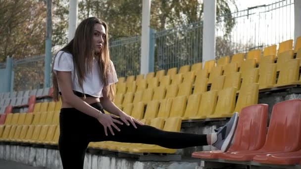 スタジアムのトライブーンでスポーツウェアを着た若い女性が足を伸ばしていて — ストック動画