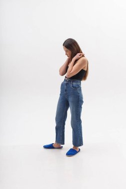 Siyah bluzlu, uzun saçlı, beyaz stüdyo arka planında izole edilmiş mavi kotlu çekici bir Asyalı kadın. Sıska, güzel bir kadın parmak arası terliklerle siklonamada poz veriyor. Güzel bayanın model testleri