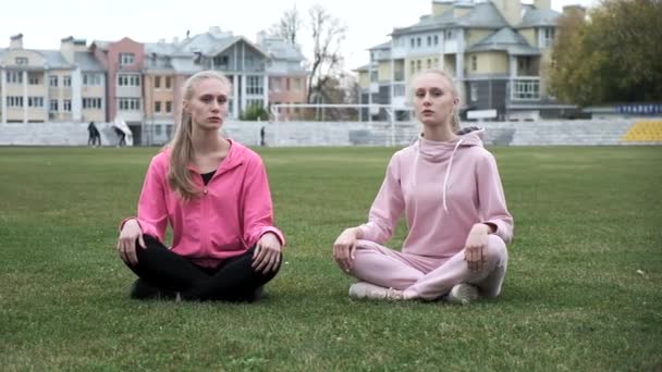Två unga passform tvillingsystrar i rosa sportkläder sitta på gräsplan i Lotus pose — Stockvideo