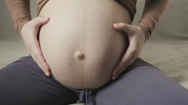 Молода вагітна жінка погладжує великий живіт на дивані, роблячи форму серця руками — стокове відео