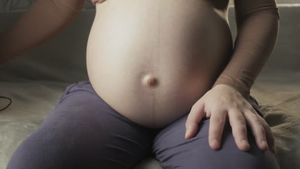 Jovem grávida sentada no sofá dentro de casa, aplicando fones de ouvido em sua barriga — Vídeo de Stock