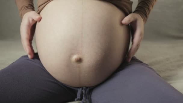 Молодая беременная женщина гладит большой живот на диване, создавая форму сердца руками — стоковое видео