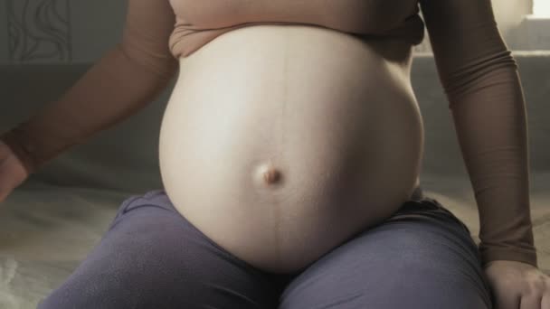 Jonge zwangere vrouw zitten op de bank binnen, het aanbrengen van koptelefoon op haar buik — Stockvideo