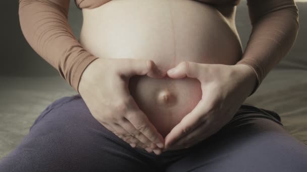 Молодая беременная женщина гладит большой живот на диване, создавая форму сердца руками — стоковое видео