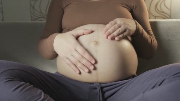 Молода вагітна жінка погладжує великий живіт на дивані, роблячи форму серця руками — стокове відео