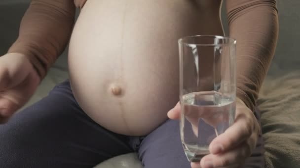 Young pregnant woman sitting on couch, taking medicine, drinking glass of water — Stockvideo