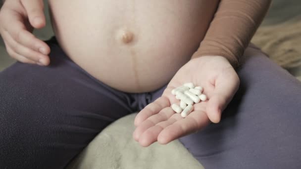 Беременная женщина сидит на диване, держит кучу таблеток в ладони, гладит живот — стоковое видео
