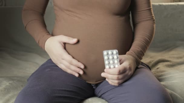 Pregnant woman sitting on couch, holding package of pills in palm, strokes belly — Video