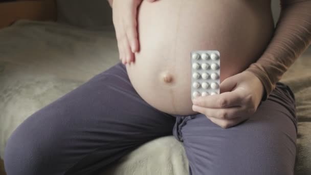 Pregnant woman sitting on couch, holding package of pills in palm, strokes belly — Stockvideo