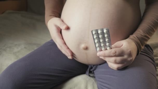 Беременная женщина сидит на диване, держит пакет таблеток в ладони, гладит живот — стоковое видео