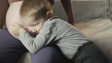 Kanepede oturan hamile kadın, küçük çocuk karnındaki bebek hareketlerini dinler.