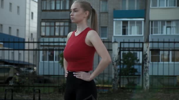 Junge, durchtrainierte blonde, vollbusige Frau mit Pferdeschwanz in rotem Top atmet am Sportplatz aus — Stockvideo