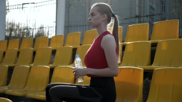 Sportliches hübsches Weibchen sitzt auf der Tribüne, trinkt Wasser aus Plastikflasche — Stockvideo