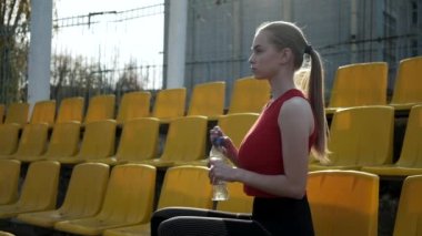 Sportif, güzel bir kadın stad tribününde oturuyor, plastik şişeden su içiyor.