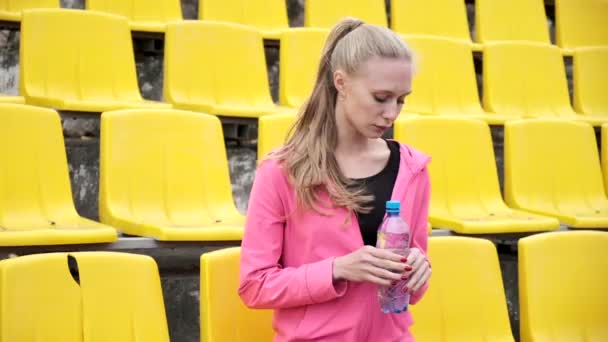 Sportovní hezká fena sedí na staduim tribune, drží plastovou láhev vody — Stock video