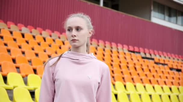 Młoda kobieta w odzieży sportowej stojąca na tle kolorowych foteli stadionowych — Wideo stockowe