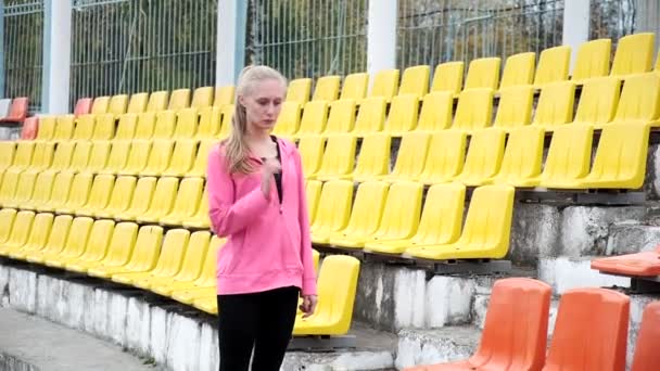 Ung kvinna i sportkläder stående på bakgrund av färgglada stadion säten — Stockvideo