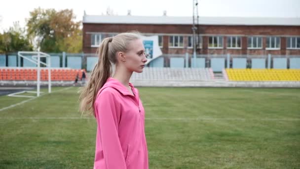 Młoda kobieta w odzieży sportowej stojąca na tle zielonego trawnika boiska do piłki nożnej — Wideo stockowe
