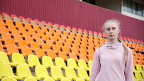 Junge Frau in Sportkleidung steht vor dem Hintergrund bunter Stadionsitze — Stockvideo