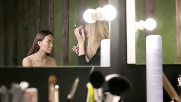 Maquillaje artista haciendo maquillaje para chica asiática en salón de belleza, reflexión en espejo — Vídeo de stock