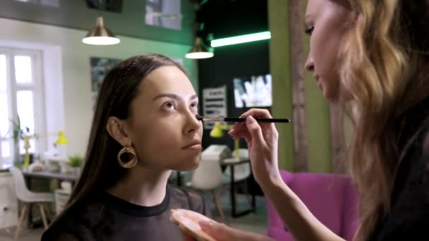 Artista de maquillaje aplica crema de tono base en la cara de las niñas usando cepillo en el salón de belleza — Vídeo de stock