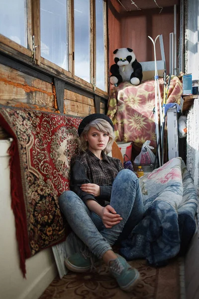 Genç, beyaz, güzel bir kadın. Balkonda oturuyor. Karışıklık ve farklı ıvır zıvırlar arasında.