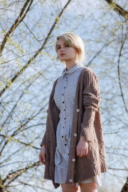 Ağaçların önünde benekli elbise ve süveter giyen beyaz bir kadın.