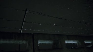 Soğuk, karanlık kış gecesinde, kar yağarken beton çitin üzerinden dikenli teller örülmüş.