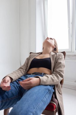 Stüdyoda çenesi yukarıda, sandalyeye uzanmış uzun saçlı beyaz bir kadın.